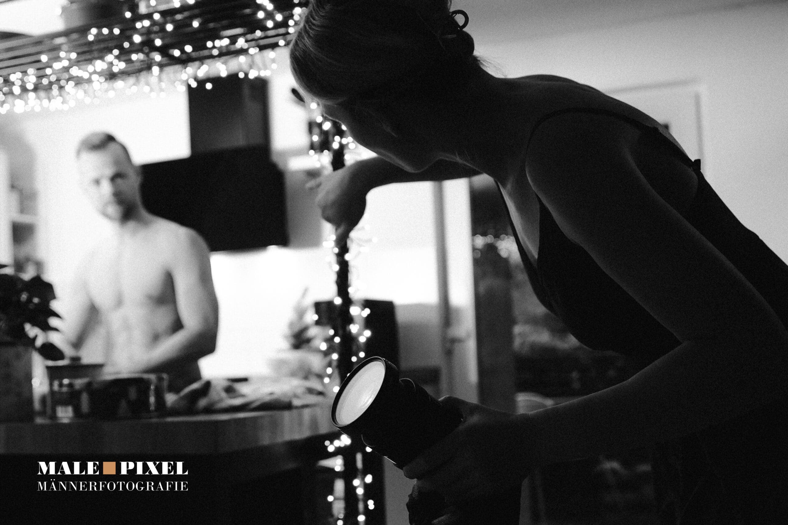 Fotografin arbeitet mit Kamera und Licht in einer Wohnungsszene, während ein oberkörperfreier Mann im Hintergrund steht, Schwarzweißaufnahme.