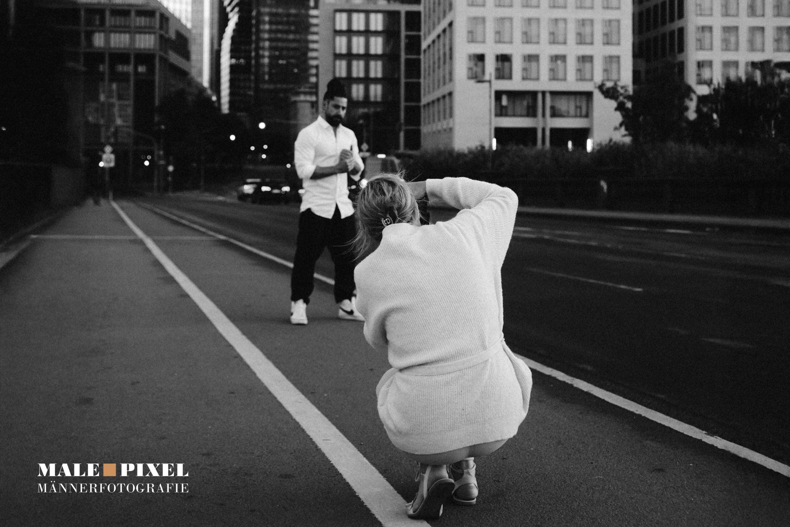 Fotografin kniet auf einer Straße und fotografiert einen Mann vor urbaner Kulisse in Schwarzweiß.