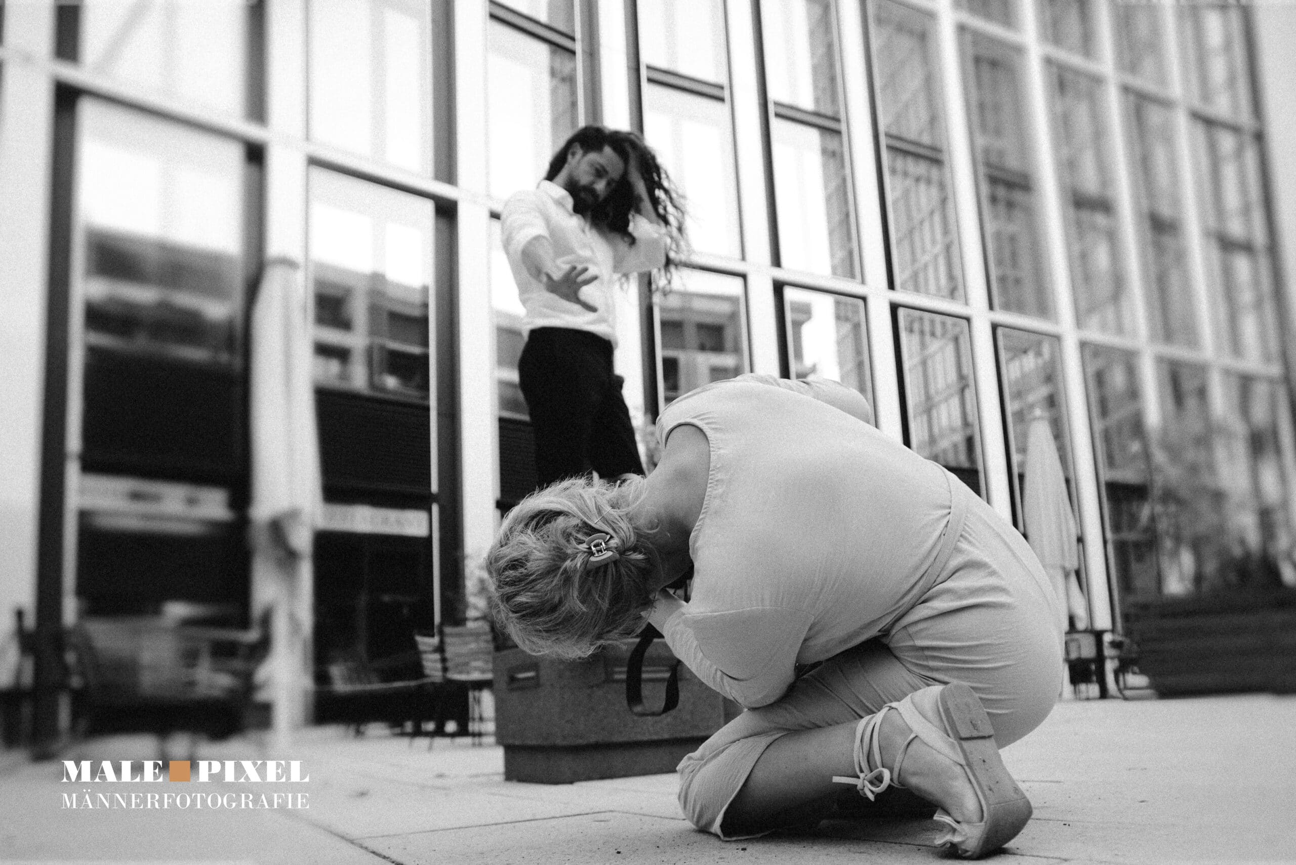 Fotografin kniet auf dem Boden und fotografiert einen Mann, der vor einer Glasfassade posiert, Schwarzweißaufnahme.