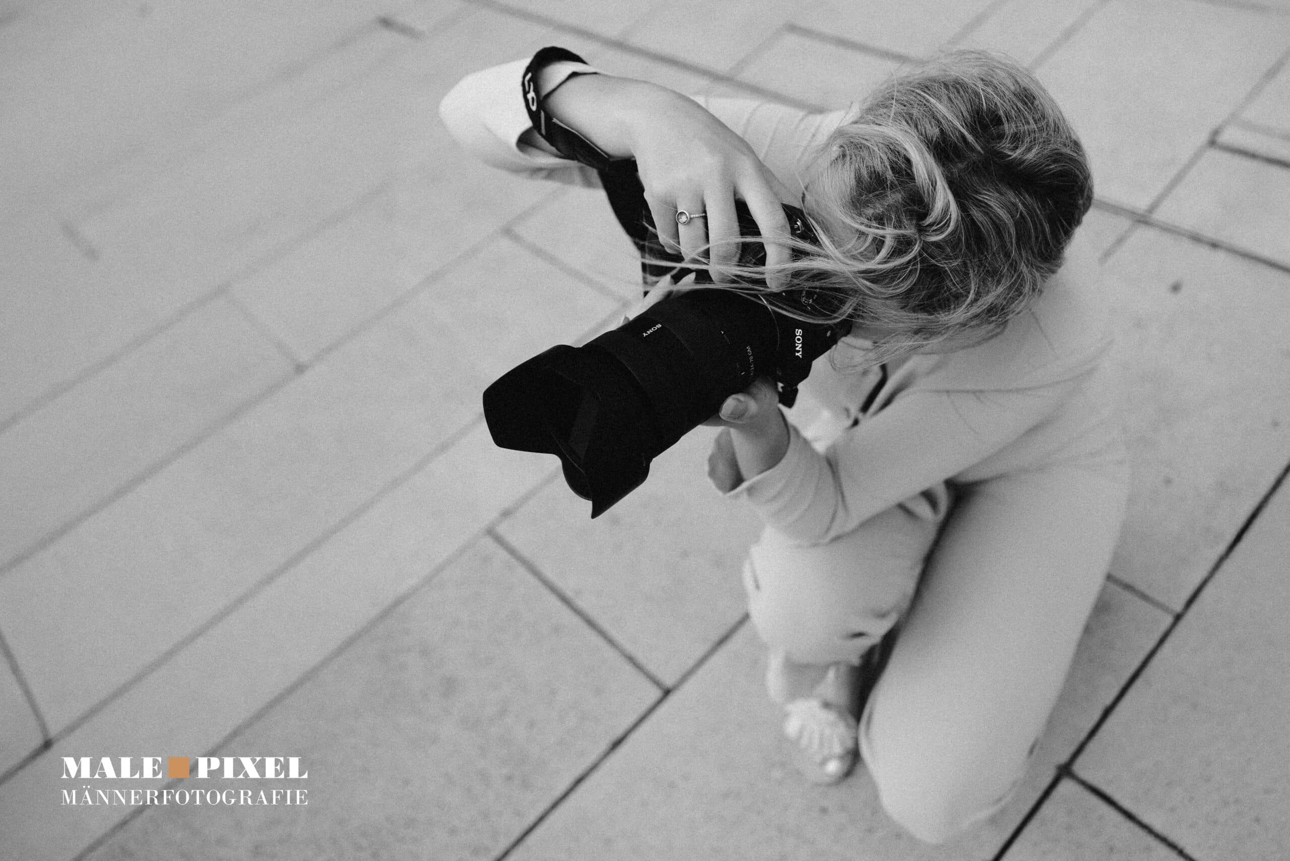 Fotografin kniet auf dem Boden und fotografiert mit einer Kamera in einer Schwarzweißaufnahme von oben.