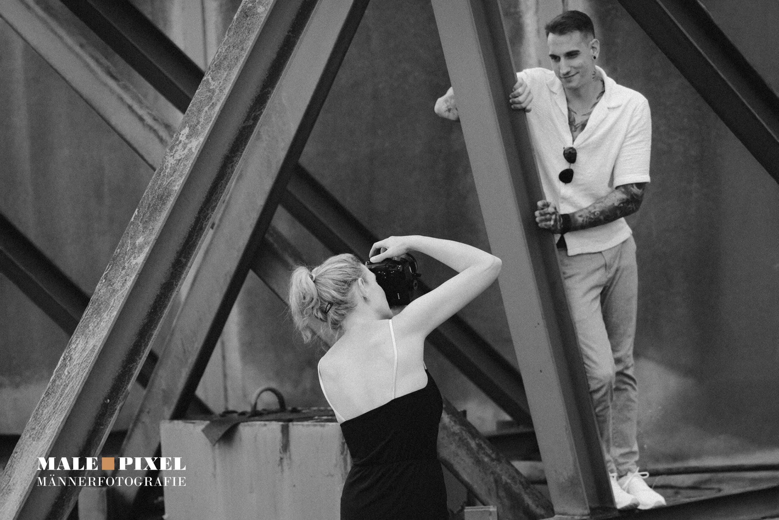 Fotografin fotografiert einen tätowierten Mann in einer industriellen Umgebung, Schwarzweißaufnahme.