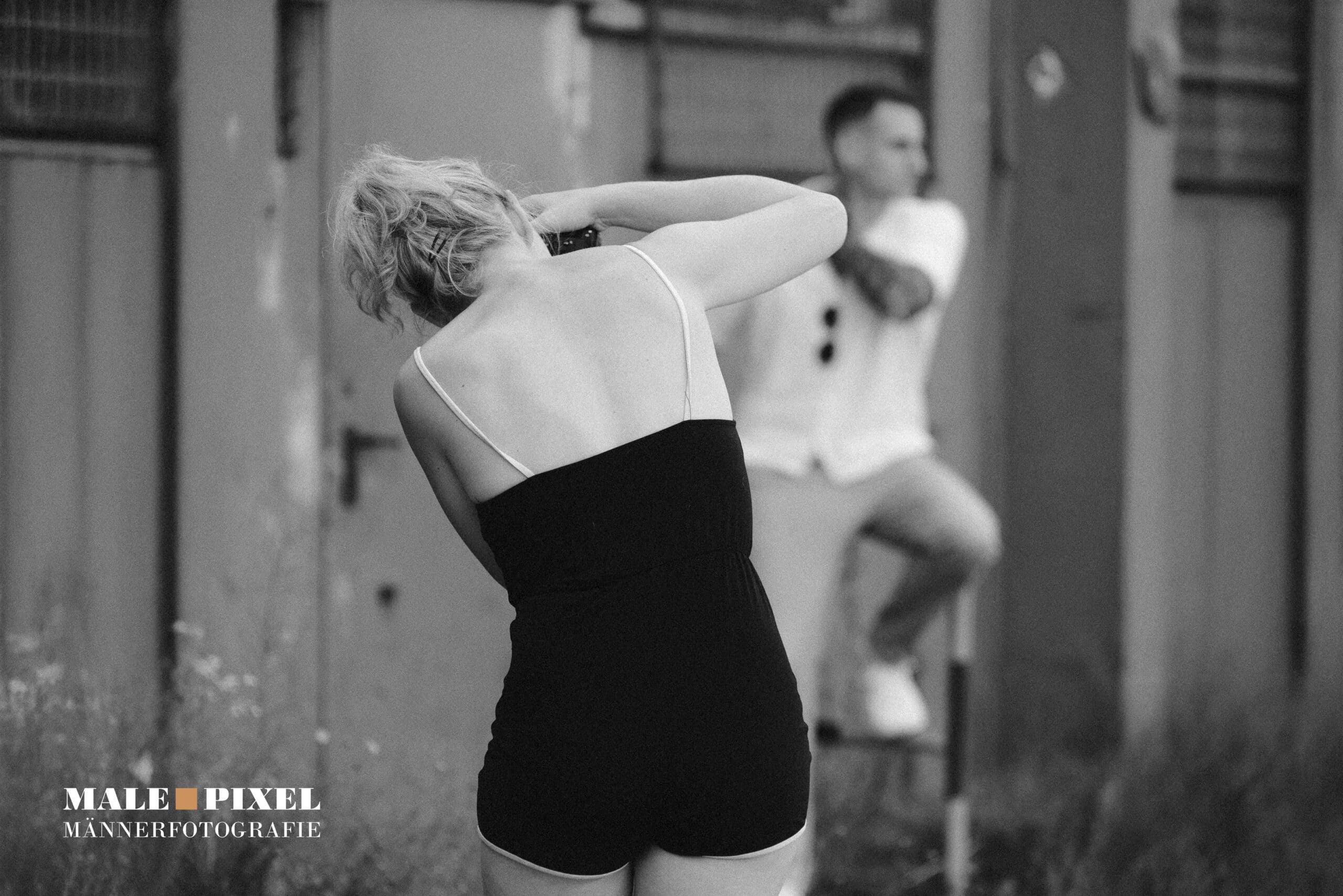 Fotografin steht in einem schwarzen Outfit und fotografiert einen sitzenden Mann in urbaner Umgebung, Schwarzweißaufnahme.