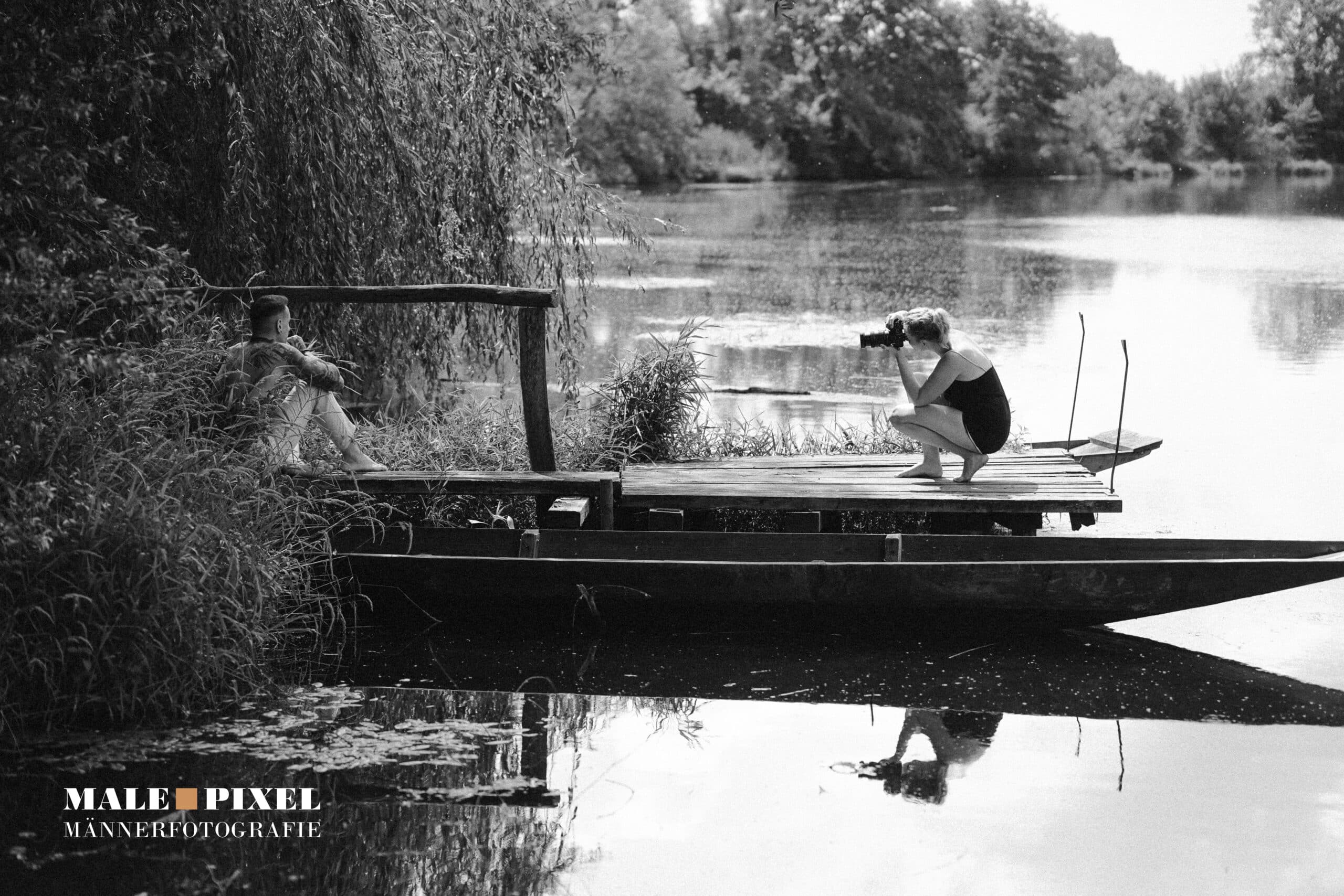 Fotografin hockt auf einem Steg und fotografiert einen Mann, der im Schilf am See sitzt, Schwarzweißaufnahme.