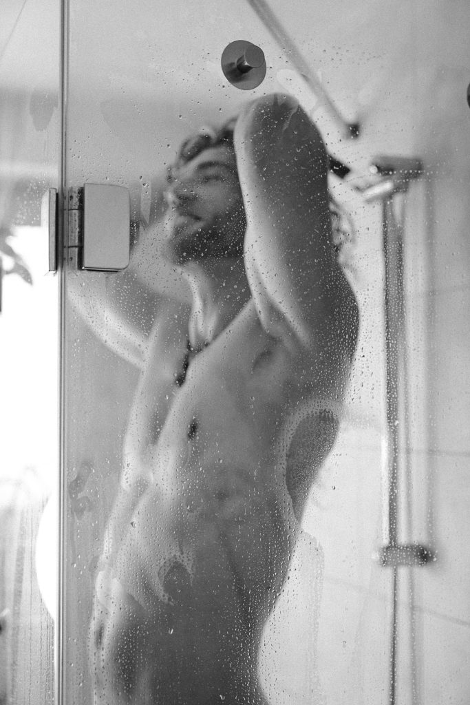 Künstlerische Schwarzweiß-Aktfotografie eines nackten Mannes hinter einer mit Wassertropfen bedeckten Duschscheibe, die Arme über dem Kopf.