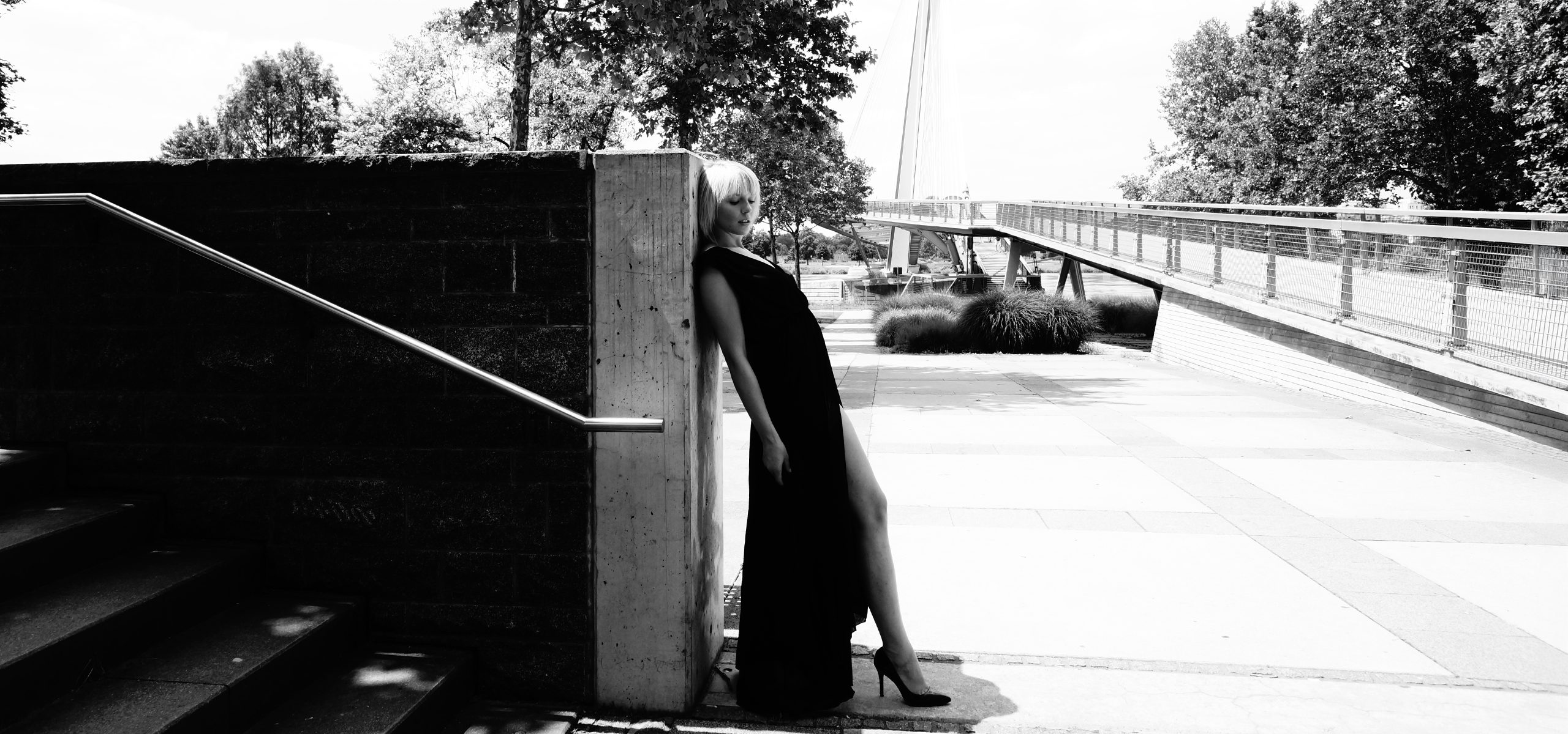 Schwarzweißaufnahme der Fotografin Natalia Schmalz in einem langen schwarzen Kleid, angelehnt an eine Betonwand im urbanen Außenbereich.