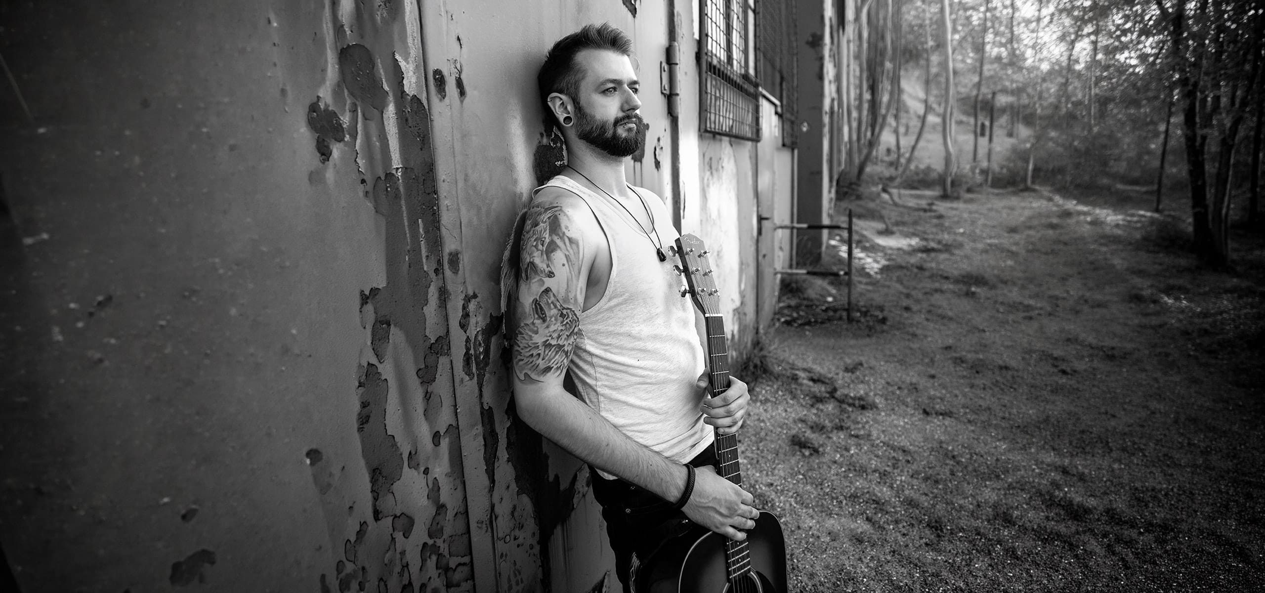 Tätowierter Mann mit Gitarre lehnt an alter Industrie-Wand – Schwarzweiß Männerportrait