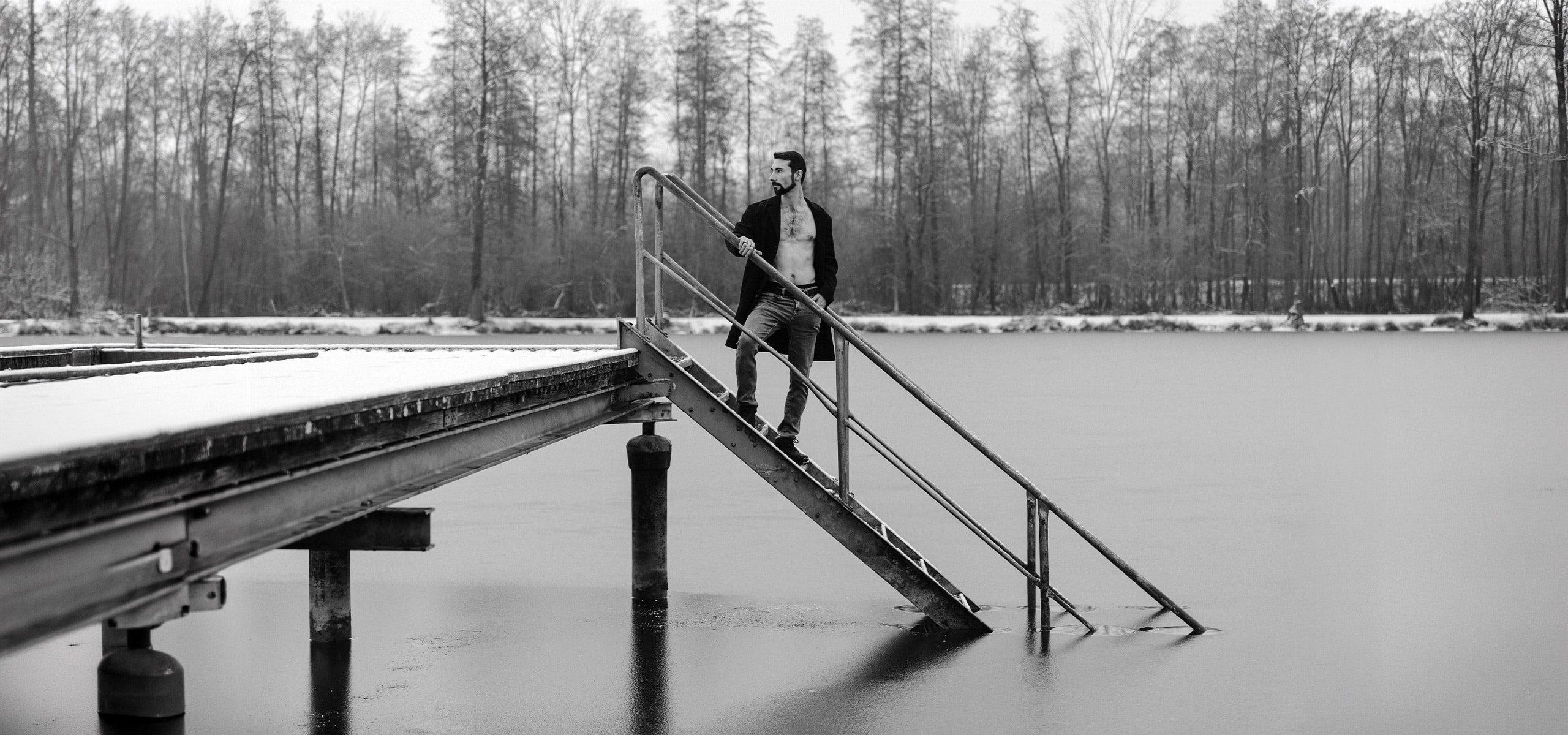 Mann steht auf Treppe eines Stegs am See – Schwarzweiß Männerportrait im Winter